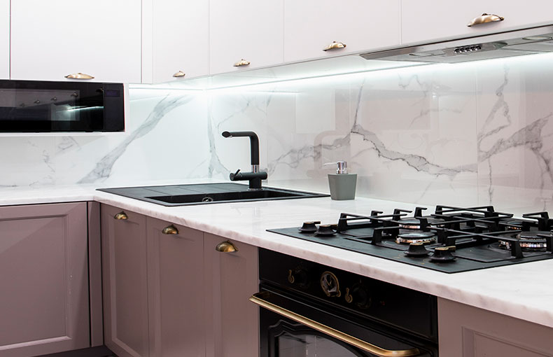 Cucina moderna con alzatina in marmo, mobili bianchi, piano cottura a gas da incasso, rubinetto nero, forno e illuminazione sotto i mobili, che creano un’atmosfera pulita ed elegante.