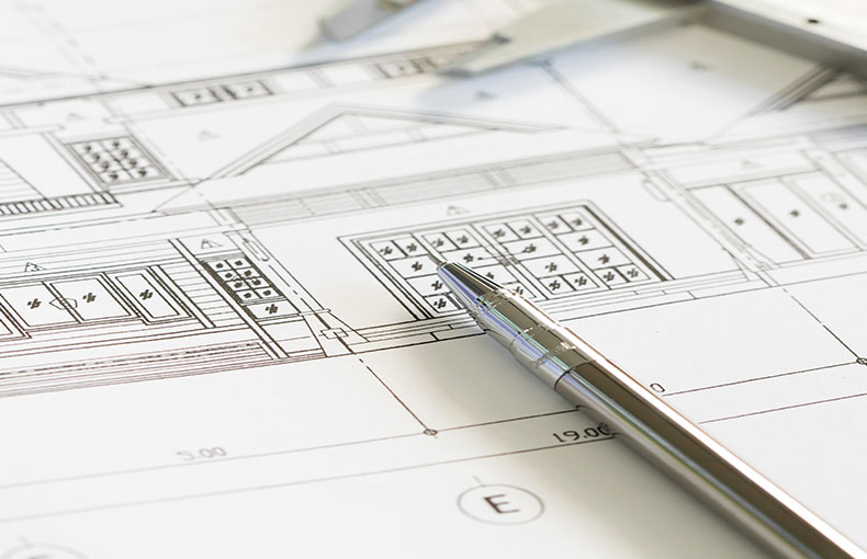 Un primo piano di una penna argentata appoggiata su progetti architettonici, che mostrano disegni dettagliati dell'edificio, misure ed elementi di progettazione. Sullo sfondo sono visibili gli strumenti da disegno.