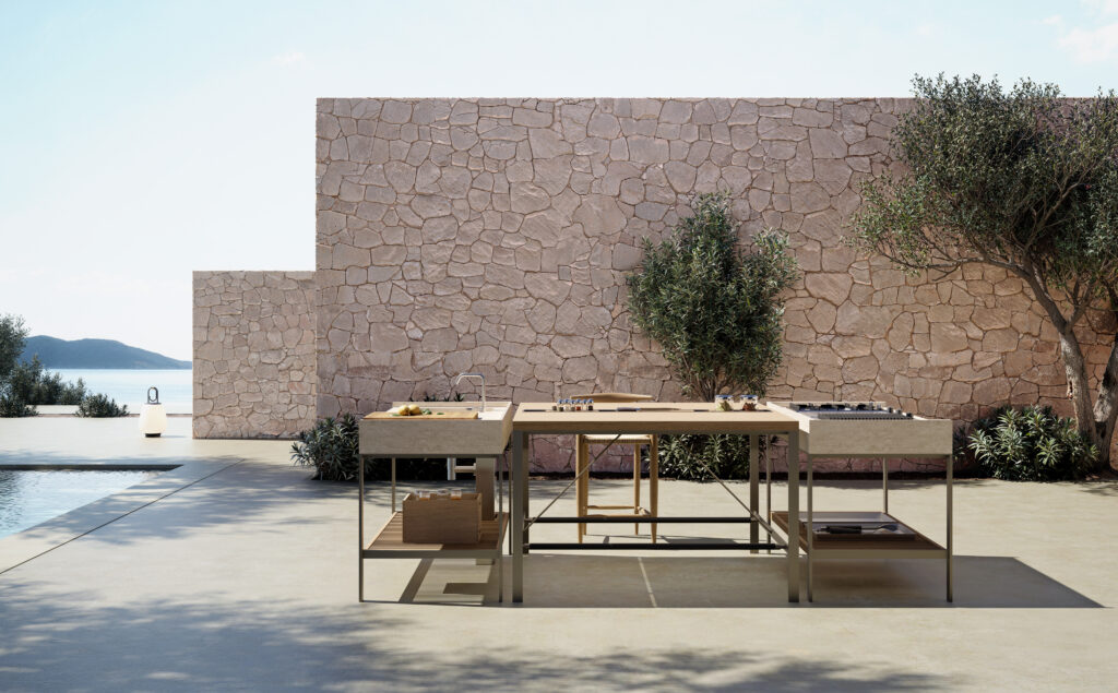 Moderna cucina all'aperto con tavoli in metallo, piano di lavoro in legno, parete in pietra e vista sulla piscina e sugli ulivi.