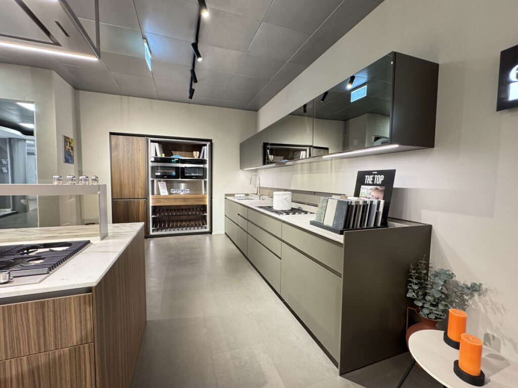 Cucina moderna con isola centrale, mobili lineari, piano di lavoro lungo, elettrodomestici da incasso e tonalità neutre.
