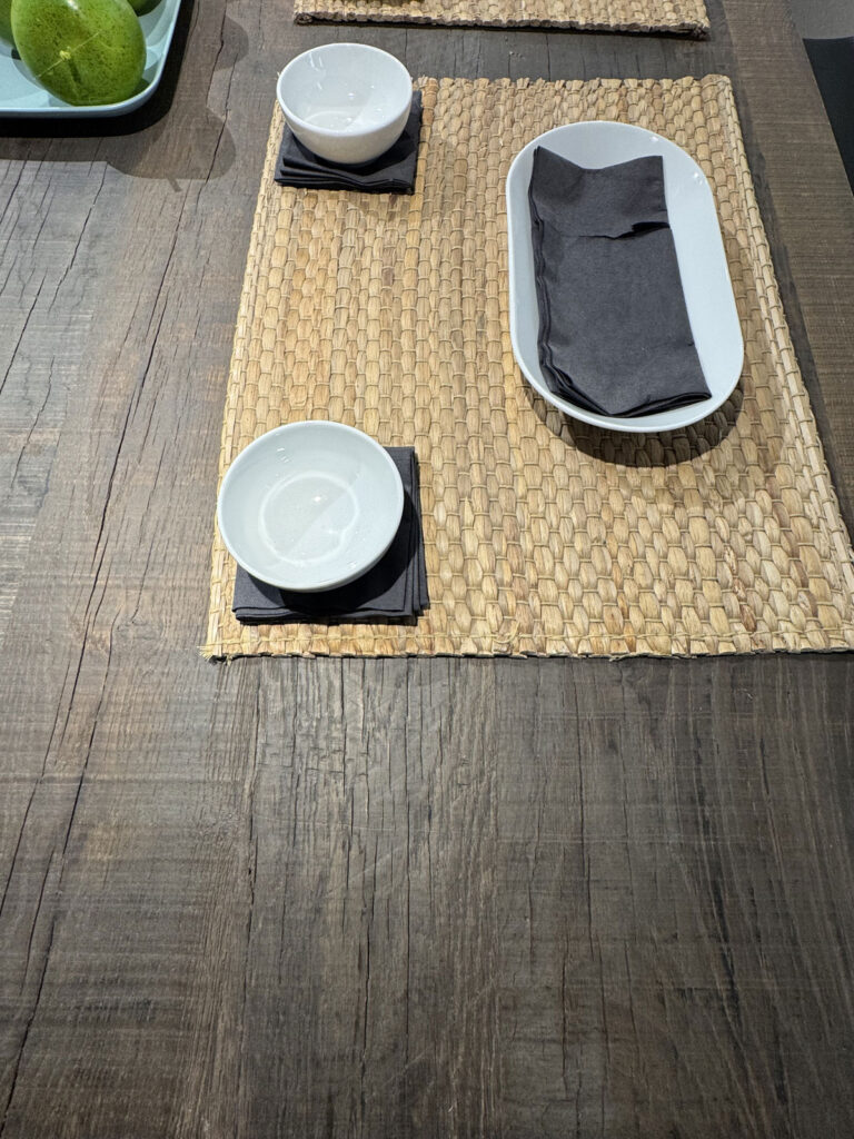 Tavolo in legno con tovaglietta intrecciata, piatto bianco ovale, due ciotole e un vassoio di mele verdi.