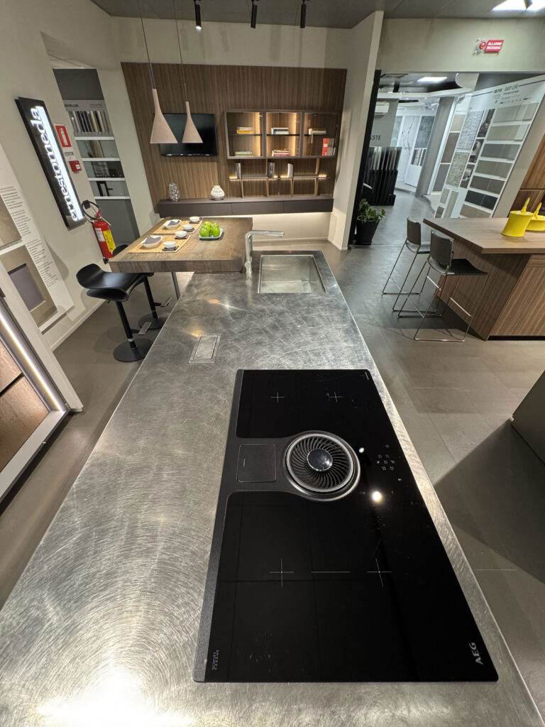 Showroom di cucine moderne Arredo 3 con isola in acciaio inossidabile, piano cottura a induzione, sgabelli e eleganti lampade a sospensione.