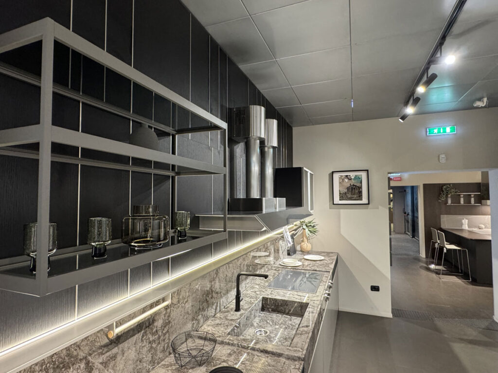 Cucina moderna K-Lab Ernestomeda con mobili scuri, pensili aperti e piano di lavoro in marmo, collegata a un altro ambiente.