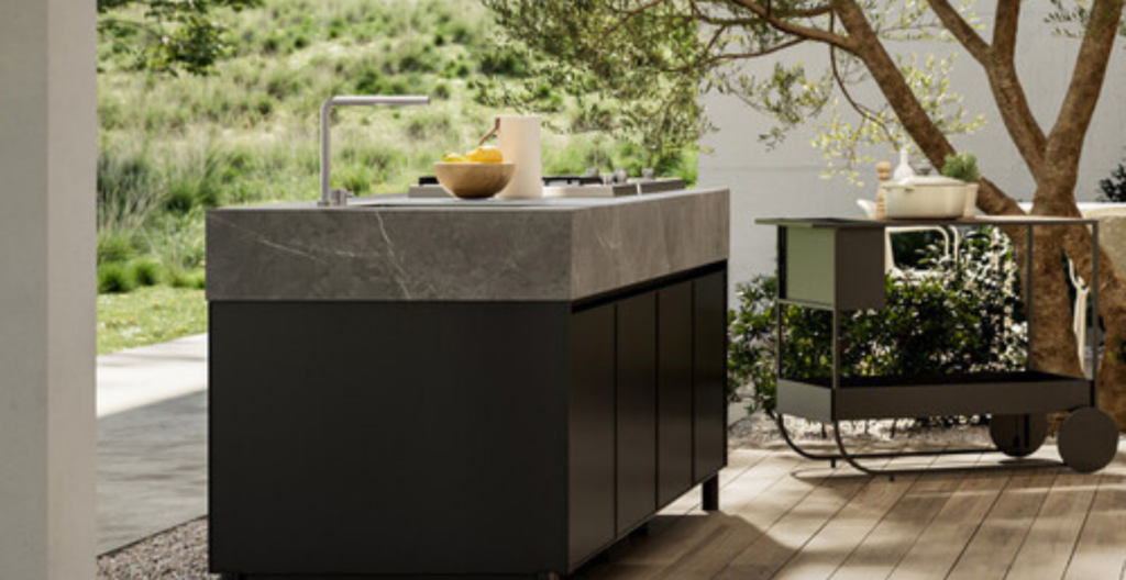 Moderna cucina all'aperto con isola nera, piano di lavoro in pietra grigia e pavimento in legno, immersa nel verde.
