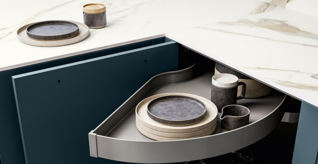 Angolo cucina moderno con base angolare estraibile, piano di lavoro bianco venato e ante blu. Piatti e tazze ben sistemati.