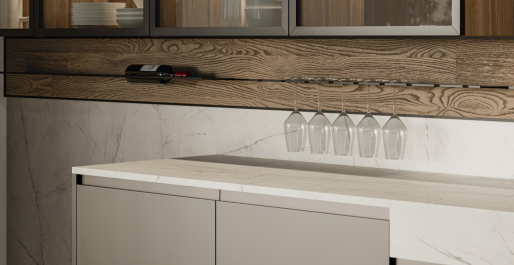 Cucina moderna con piano in marmo, mobili bassi grigi e pannello posteriore in legno. Pensili in vetro con bottiglie e piatti.