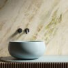 Lavabo rotondo azzurro opaco su mobile in legno rigato, rubinetti neri e piastrelle effetto marmo Del Conca.