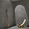 Pareti e pavimento in grigio Stone Edition di Del Conca, sedia scultorea beige, luce naturale in un ambiente minimalista.