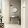 Bagno moderno con piastrelle beige e grigie della Ceramica Del Conca, lavabo piccolo, specchio rotondo e luce naturale.
